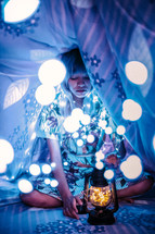 a young woman holding fairy lights and a lantern under sheets 