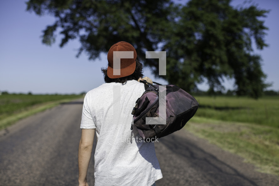 man standing in the middle of a road with his back to the camera carrying a bag