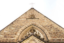 Looking up at the gable roof of a Gothic-style church. A white cross sits at the head of the roof's ridge.