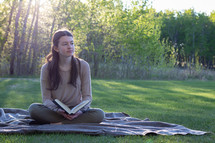 a girl sitting on a picnic blanket reading a Bible 