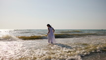 Woman in a white dress on a beach standing in shallow water.
