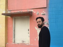 a man with a beard standing in front of a multi colored wall 