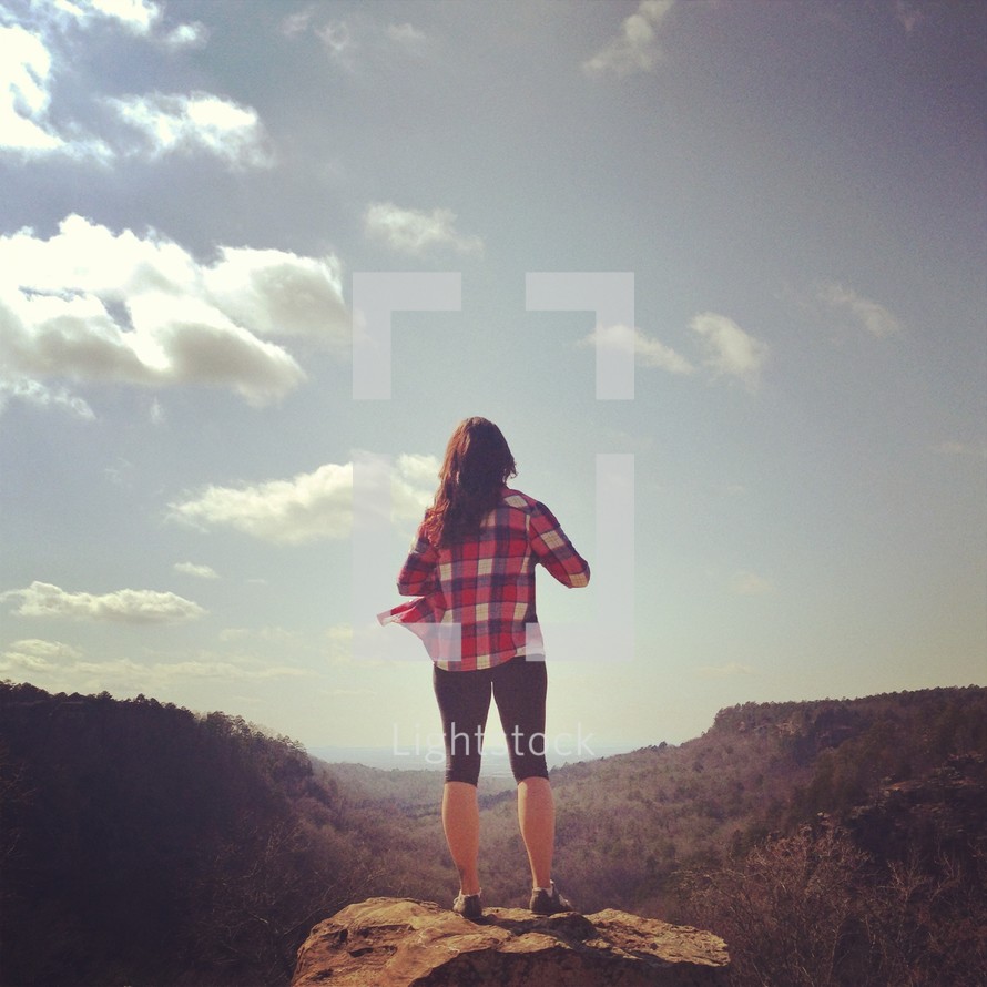 woman standing on a rock at a mountain's edge looking up a the sky