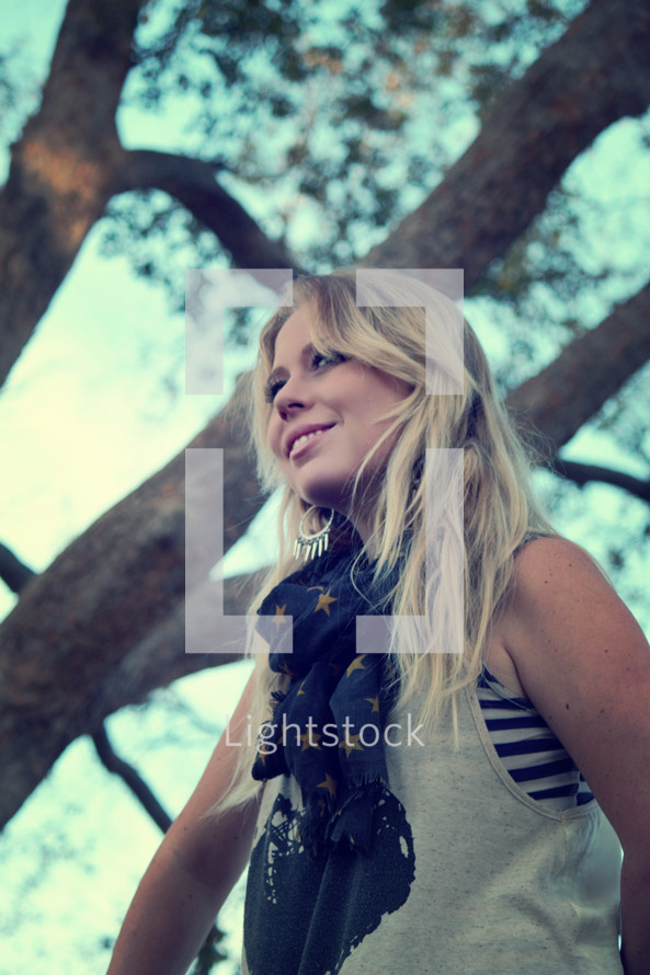 blonde woman wearing a scarf standing in front of a tree smiling 