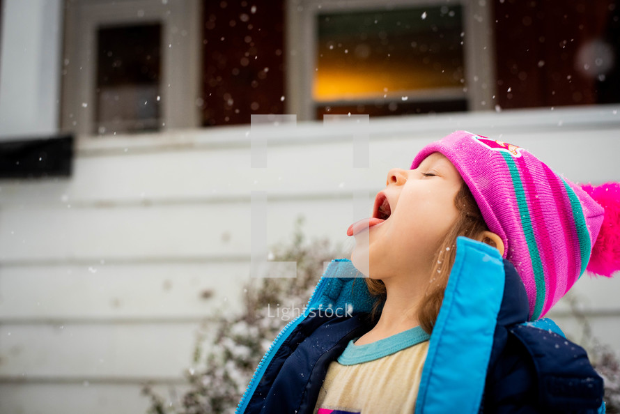 A little girl catching snow on her tongue while wearing winter clothes outside. 
