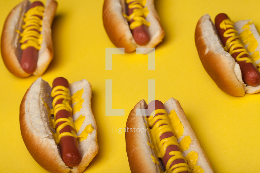 Hot dogs and buns drizzled with mustard on a bright yellow background.