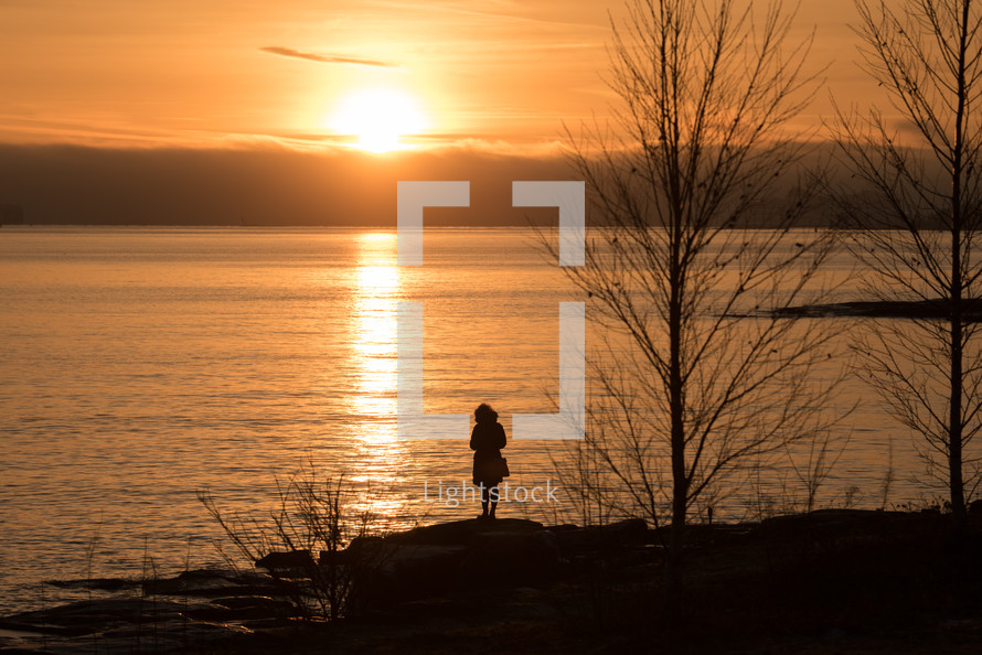 silhouette standing on a lake shore at sunset and glow of sunlight in the shape of a cross