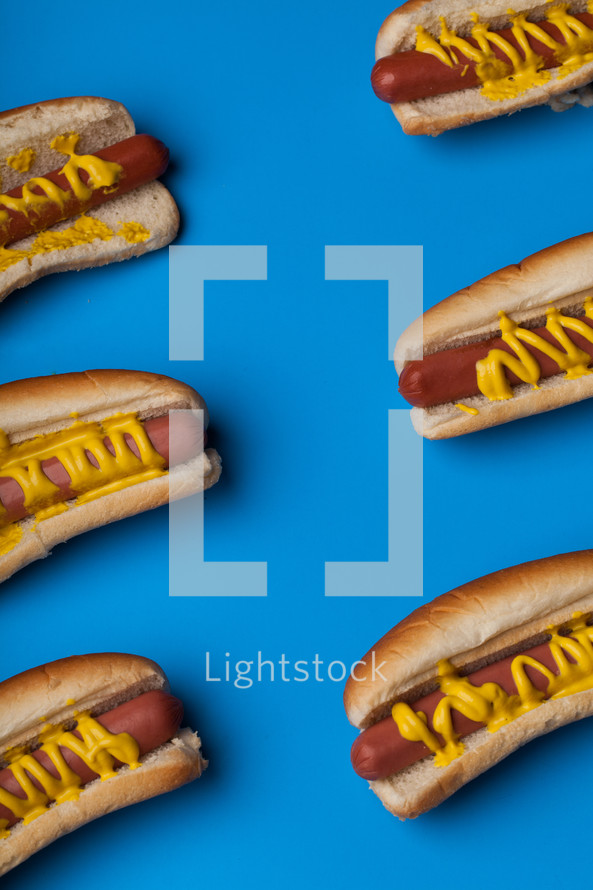 Hot dogs and buns drizzled with mustard on a bright blue background.