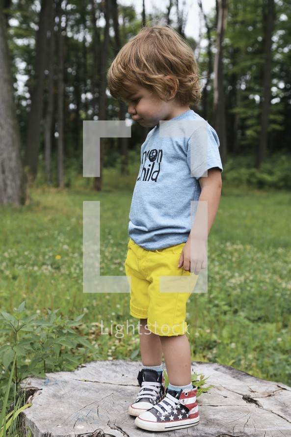 a toddler boy standing outdoors looking down at the ground 