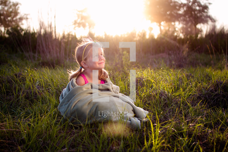 a little girl wrapped in a blanket sitting in tall grasses at sunset 