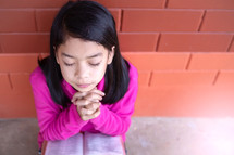 a girl praying in front of a brick wall 