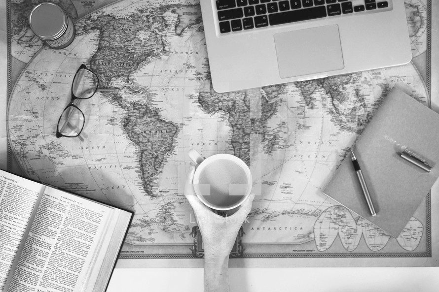 open Bible, mug, reading glasses, laptop, pen, journal, and ink on a world map 