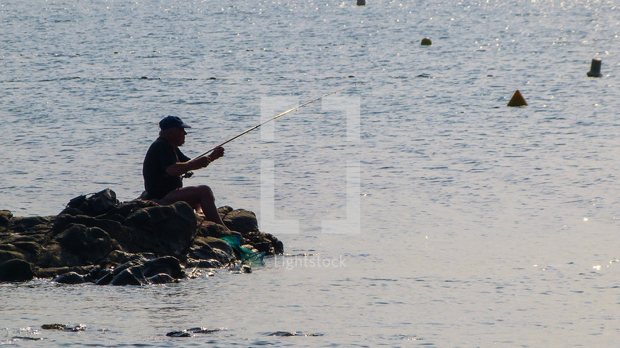 a man fishing at the ocean sitting on rocks 