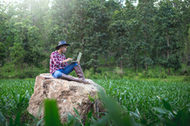 Young farmer sitting on a big stone and checking his corn field and working on laptop computer,