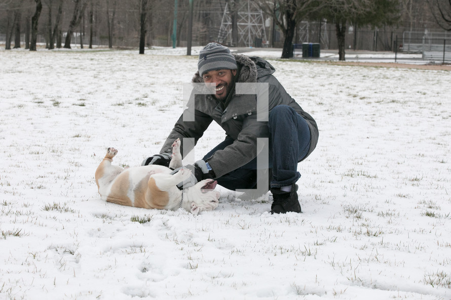 a smiling man playing in snow with a bulldog 