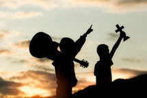 silhouettes of two boys standing outdoors at sunset holding a guitar and cross 