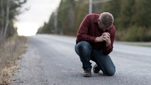 a man kneeling in prayer in the middle of a rural road 