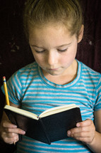 A girl reading a notebook and holding a pencil.