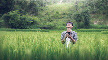 a young man standing in a rice field holding a cellphone praying to God 