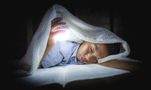 a little boy reading a Bible under a blanket holding a light