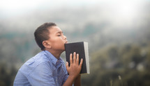 a boy holding a Bible praying outdoors 
