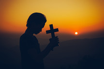 Young man praying and holding Cross at sunset 