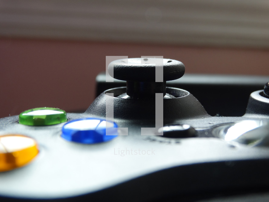 Close up of analog thumb stick on black gaming controller with blue, green and yellow buttons shining in the light; game night concept