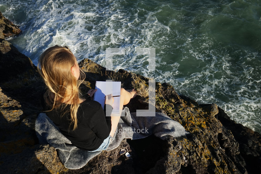 woman sitting on rocks along a shore writing in a journal 