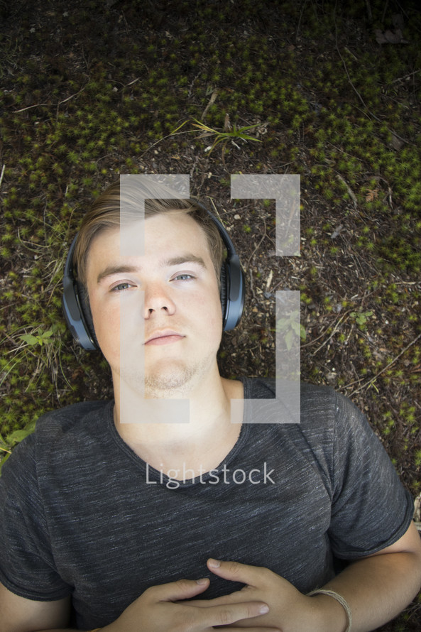 Teenager listening to music and lying down on grass and earthy background looking at camera
