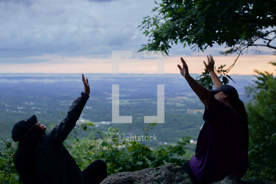 friends with raised hands sitting at the edge of a cliff on a mountaintop 