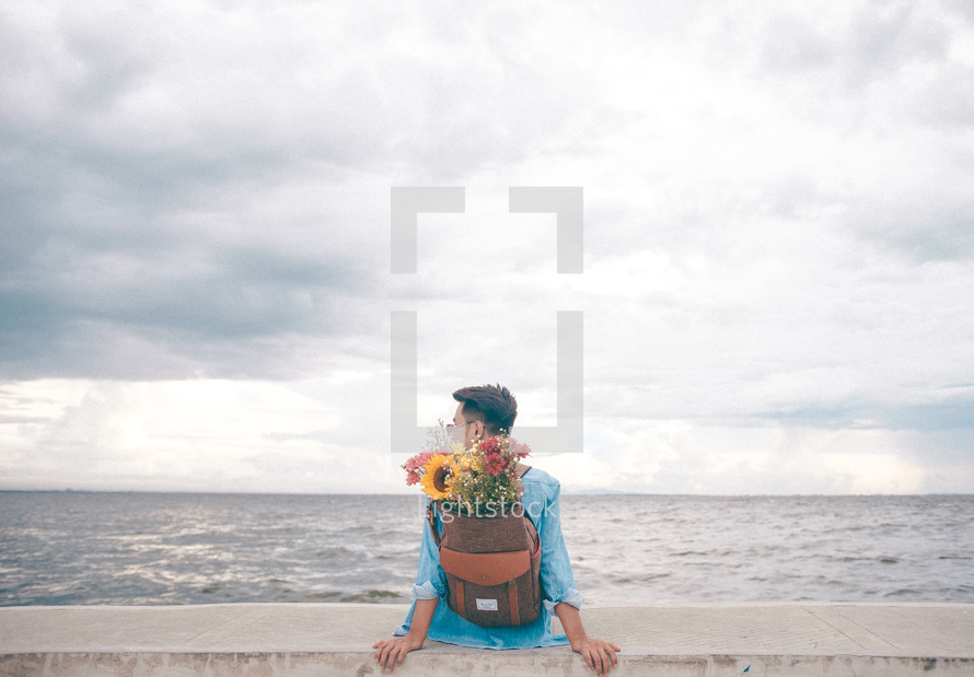 a man with flowers in a backpack sitting at a shore 
