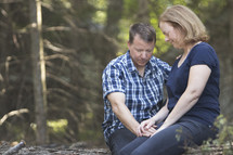 Couple praying together in wooded area and holding hands while sitting on log