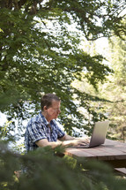 Middle aged man working on laptop computer in rural wooded environment in woods