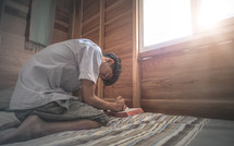 a teen kneeling on a bed by a window praying 