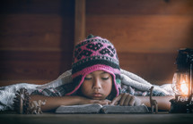 a child in a beanie reading a Bible under a blanket by a lantern 