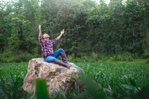 Young farmer sitting on a big stone with hands raised praising God 
