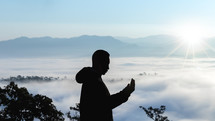 a man in a coat standing on a mountaintop in the clouds praying 