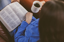 A woman siting in a chair reading her Bible and drinking coffee