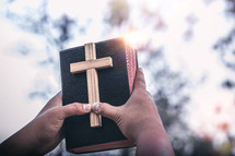 a man holding up a wooden cross and Bible outdoors 