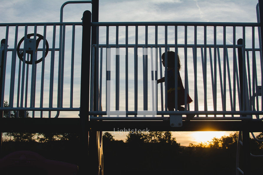 A young child (girl) runs across playground equipment with a sunset behind her.