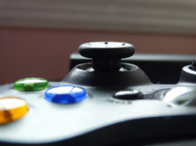 Close up of analog thumb stick on black gaming controller with blue, green and yellow buttons shining in the light; game night concept