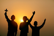 silhouettes of three children standing outdoors at sunset holding a cross