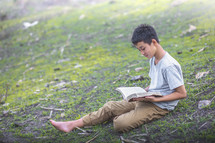 a boy sitting on a hill reading a Bible 