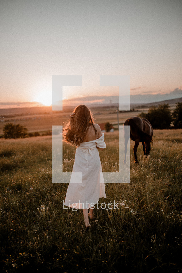 Woman in white dress walking by horse at sunset