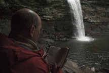 man reading a pocket Bible near a waterfall