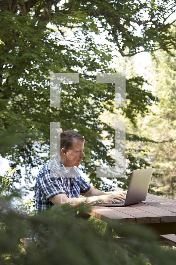Middle aged man working on laptop computer in rural wooded environment in woods
