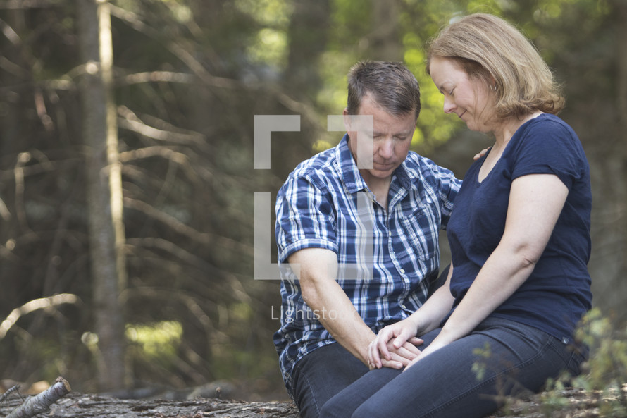Couple praying together in wooded area and holding hands while sitting on log