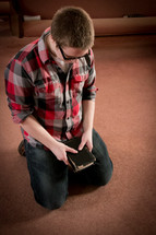 Boy kneeling in prayer, holding a Bible, in front of a church pew.