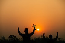 two boys holding up a cross at sunset under an orange sky 