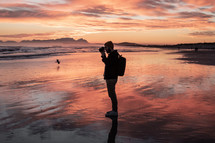 man with a camera standing on a beach at sunset 